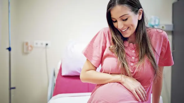 Junge Frau mit braunen Haaren hält lachend ihren Babybauch