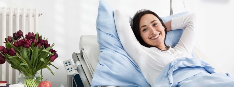Junge Frau mit schwarzen Haaren liegt lächelnd in einem Krankenhausbett