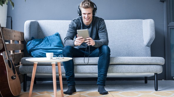 Ein junger Mann sitzt auf einem Sofa, hat Kopfhörer auf und schaut in ein Tablet