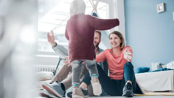 Mutter und Vater sitzen mit offenen Armen am Boden und ihr Kind läuft auf sie zu