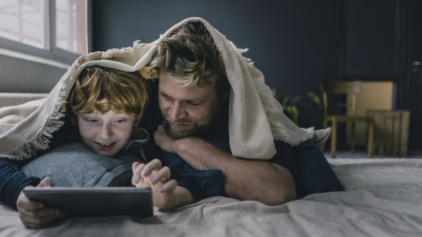 Mann und Sohn liegen unter einer Decke und blicken in ein Tablet