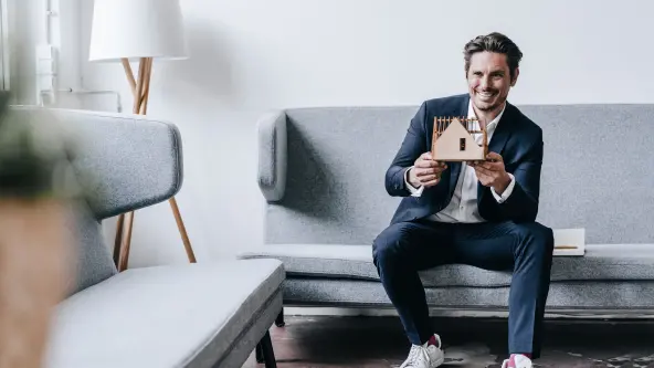Lächelnder Mann sitzt auf der Couch und hält ein Hausmodell aus Holz