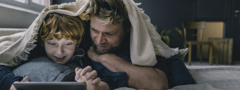 Mann und Sohn liegen unter einer Decke und blicken in ein Tablet