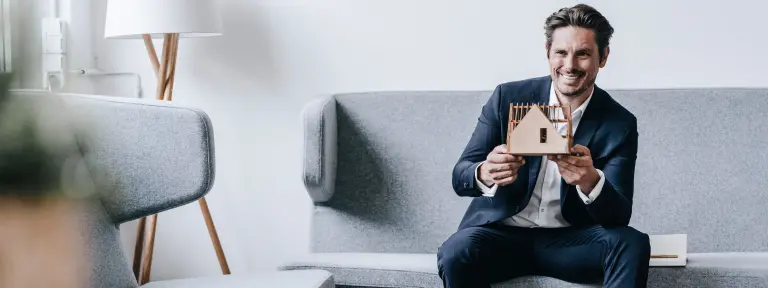 Lächelnder Mann sitzt auf der Couch und hält ein Hausmodell aus Holz