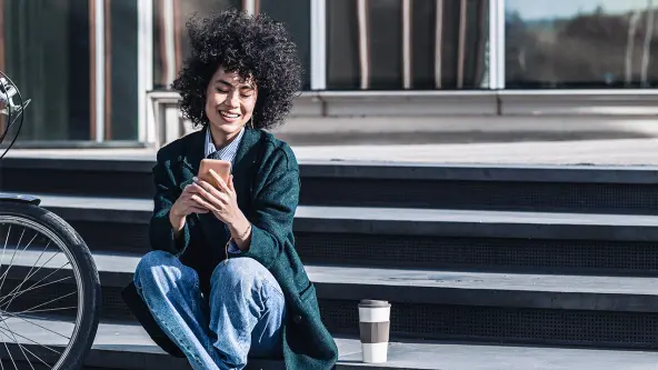 Junge Frau mit schwarzen Locken sitzt mit Handy in der Hand auf einer breiten Steintreppe