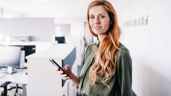Frau mit roten langen Haaren und elegantem Businessoutfit steht in einem hellen Büro und hält ein Smartphone in der Hand