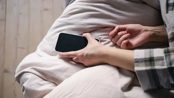 Frau liegt im Bett und greift mit der Hand nach dem Handy