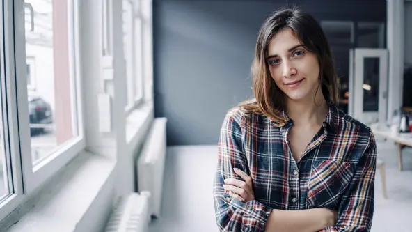 Junge Frau steht mit verschränkten Armen und einem karierten Hemd in einem hellen Raum