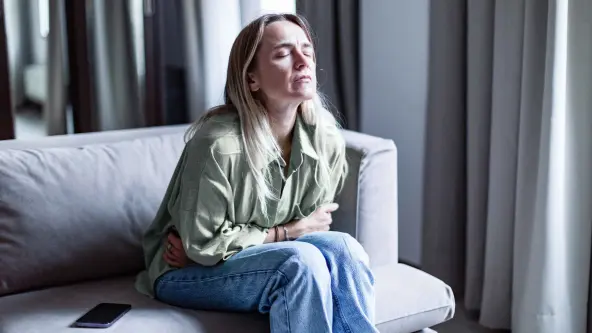 Blonde Frau sitzt gekrümmt und mit geschlossenen Augen auf einem Sofa und umklammert ihren Bauch