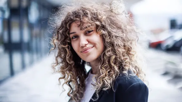 Junge Frau mit vielen Locken steht lächelnd vor einem Bürogebäude