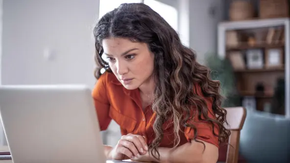 Junge Frau mit lockigen Haaren sitzt sehr tief über einen Laptop gebeugt an einem Tisch