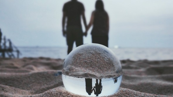 Glaskugel liegt im Sand und zeigt ein Händchen haltendes Pärchen am Strand