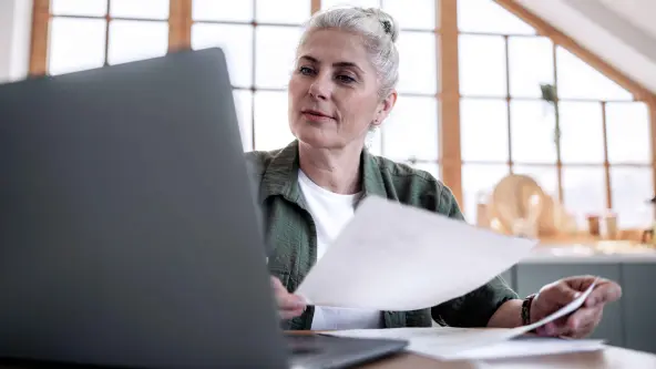 Eine ältere Frau mit weißen Haaren sitzt vor ihrem Laptop an einem Schreibtisch und überprüft ihre Post