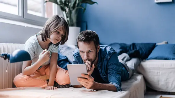 Mann liegt auf Sofa und schaut mit seinem Kind in ein Tablet
