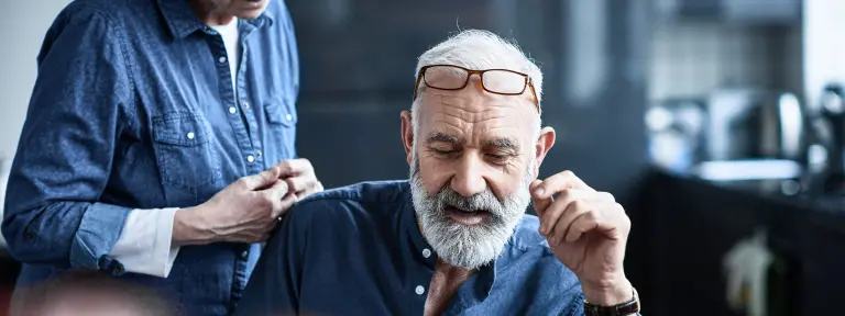 Älterer Mann mit grauem Bart und Brille auf der Stirn sitzt vor einem Laptop - neben ihm steht eine ältere Frau, die ihm über die Schulter blickt
