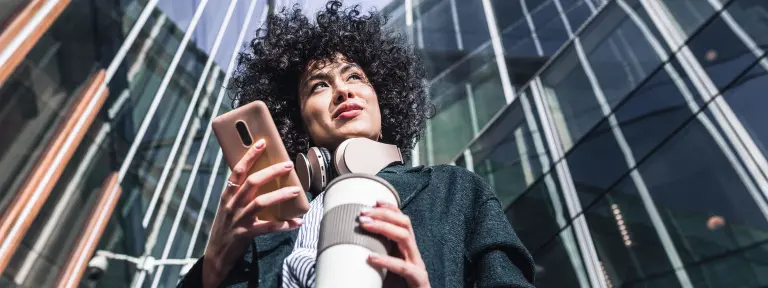 Junge Fau mit Handy und Kaffeebecher in den Händen steht vor einem großen Glasgebäude