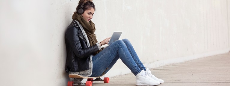 Junge Frau mit Kopfhörer sitzt auf Skateboard mit Tablet in der Hand