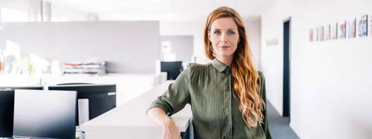 Frau mit langen roten Haaren und einem eleganten Businessoutfit steht in einem offenen und hellen Büro