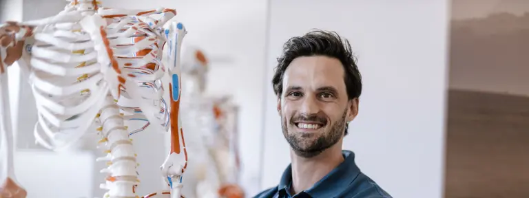 Arzt mit blauem Poloshirt und verschränkten Armen steht vor einem großen Skelettmodell