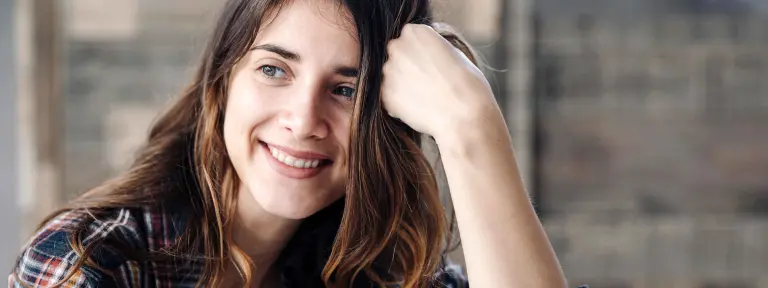 Junge Frau mit dunklem Haar stützt ihren Kopf auf einen Arm und lächelt dabei in die Kamera