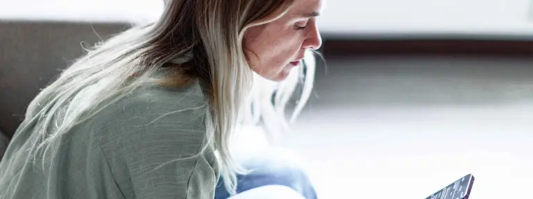 Blonde Frau sitzt auf Couch und schaut in ihr Smartphone in ihrer rechten Hand