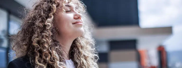 Eine junge Frau mit ganz vielen Locken atmet vor einem Bürogebäude tief ein