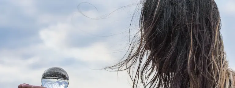 Frau mit langen Haaren hält Glaskugel in der Hand in der sich die Wolken spiegeln