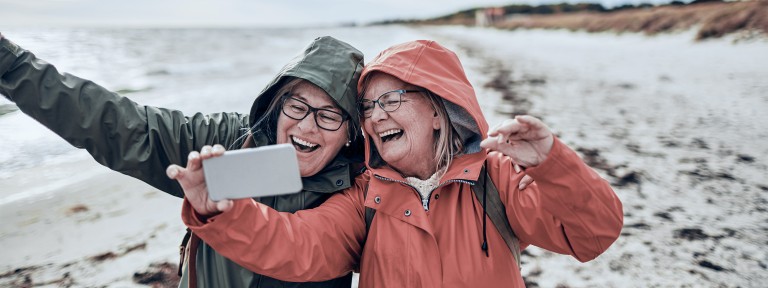 2 ältere Frauen in Regenjacken machen ein Selfie am Strand