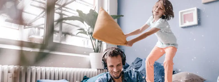 Mann sitzt mit Kopfhörer und Tablet auf Boden - während Kind hinter ihm auf der Couch mit einem Kissen wedelt