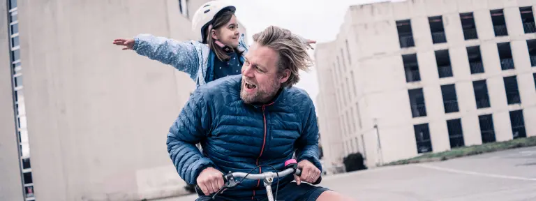 Mann sitzt auf rosafarbenen Kinderfahrrad und ein Mädchen steht hinter ihm auf dem Rad