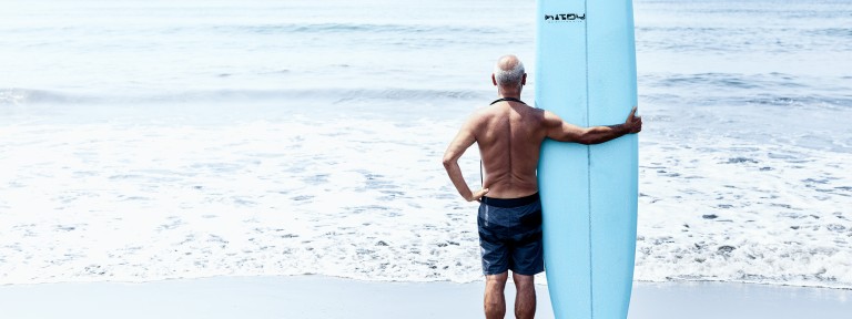 Älterer Mann steht mit einem Surfbrett am Strand und blickt ins Meer