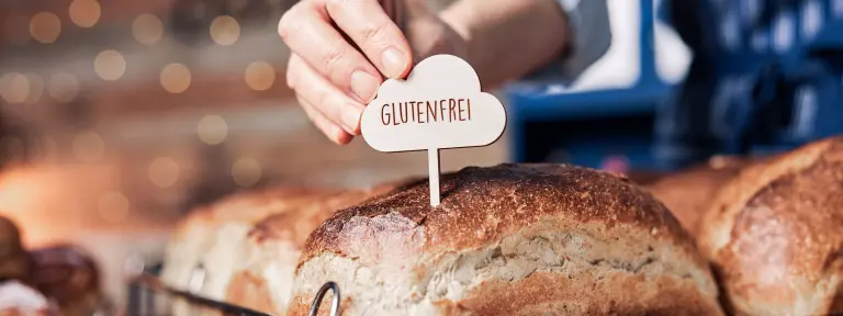 Eine Hand steckt ein Schild mit der Aufschrift "GLUTENFREI" in ein Brot bei einer Bäckerei