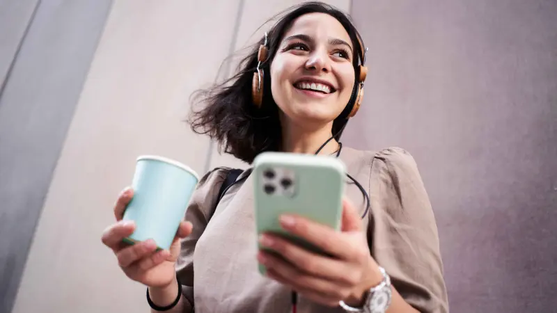 Eine junge Frau mit Kopfhörern, Kaffee und Smartphone lächelt unbeschwert