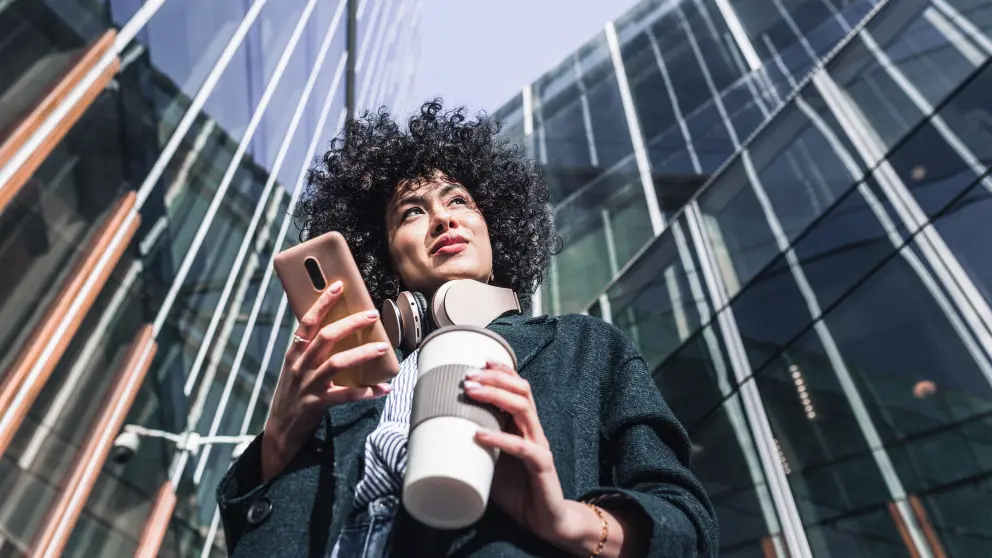 Junge Fau mit Handy und Kaffeebecher in den Händen steht vor einem großen Glasgebäude