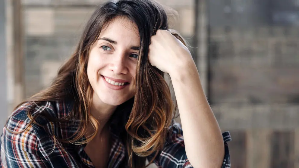 Junge Frau mit dunklem Haar stützt ihren Kopf auf einen Arm und lächelt dabei in die Kamera