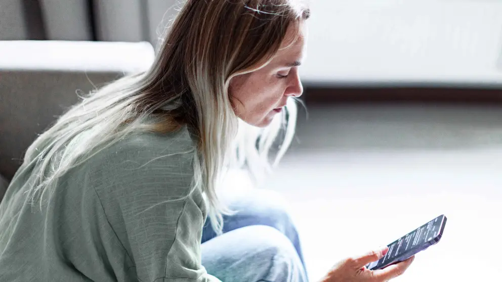 Blonde Frau sitzt auf Couch und schaut in ihr Smartphone in ihrer rechten Hand