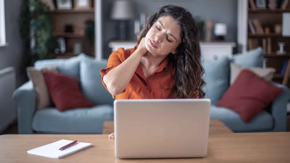 Junge Frau mit vielen Locken sitzt an einem Tisch auf dem ein Laptop steht und reibt sich ihren schmerzenden Nacken