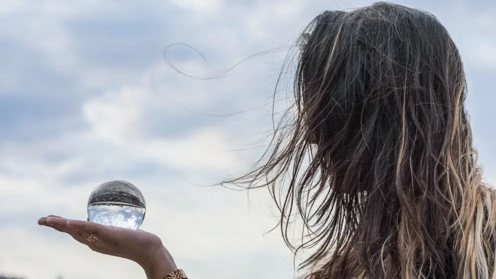 Frau mit langen Haaren hält Glaskugel in der Hand in der sich die Wolken spiegeln