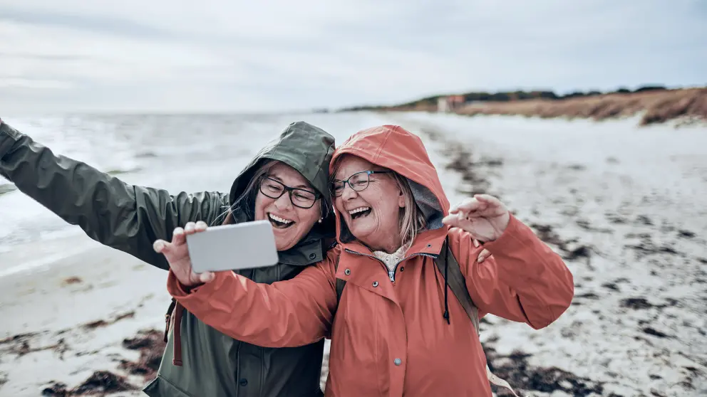 2 ältere Frauen in Regenjacken machen ein Selfie am Strand