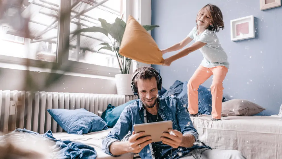 Mann sitzt mit Kopfhörer und Tablet auf Boden - während Kind hinter ihm auf der Couch mit einem Kissen wedelt