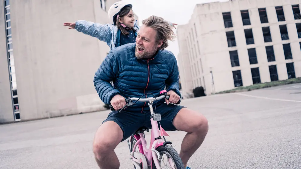 Mann sitzt auf rosafarbenen Kinderfahrrad und ein Mädchen steht hinter ihm auf dem Rad