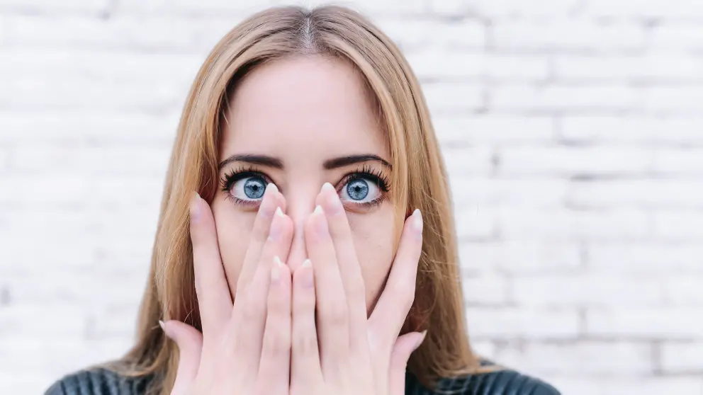 Junge Frau hält sich beide Hände vor den Mund und man sieht ihre blauen Augen