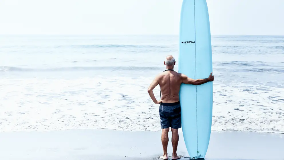 Älterer Mann steht mit einem Surfbrett am Strand und blickt ins Meer