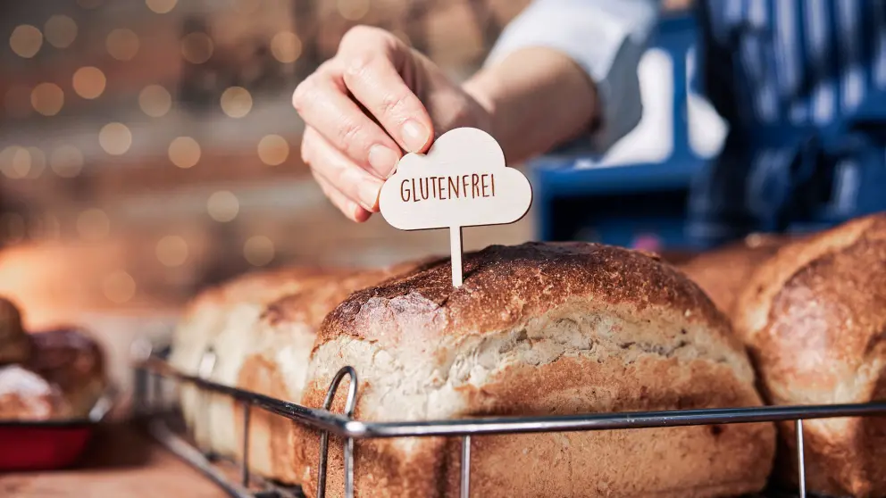 Eine Hand steckt ein Schild mit der Aufschrift "GLUTENFREI" in ein Brot bei einer Bäckerei