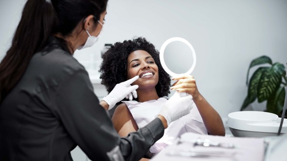 Frau mit lockigen Haaren schaut sich auf Behandlungsstuhl ihre Zähne im Spiegel an