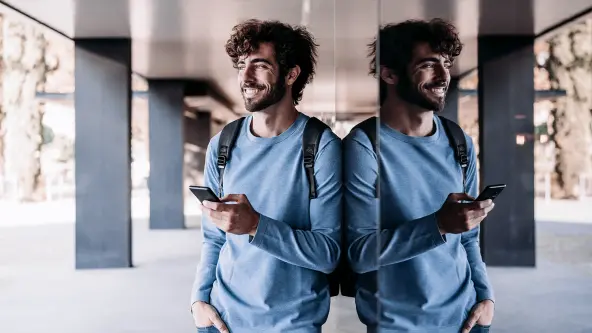 Junger Mann im blauen Pulli lehnt an Glasscheibe und hat Handy in der Hand