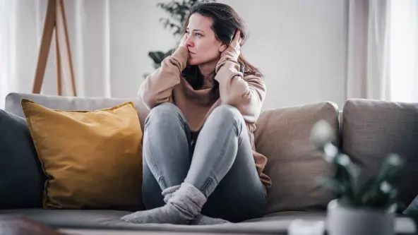 Eine Frau sitzt mit angewinkelten Beinen auf einer Couch und fährt sich mit ihren Händen durch ihre Haare 