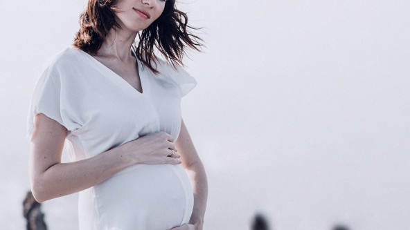 Schwangere Frau trägt weißes Kleid und hält sich ihren Bauch