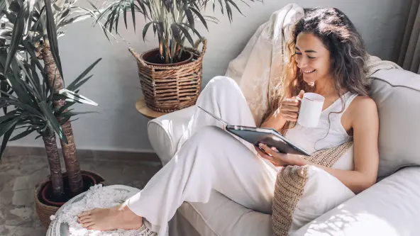 Junge Frau sitzt mit Kaffeetasse und Tablet auf einem Sessel