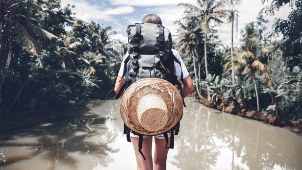 Frau läuft mit großem Rucksack am Rücken durch einen seichten Fluss im Dschungel